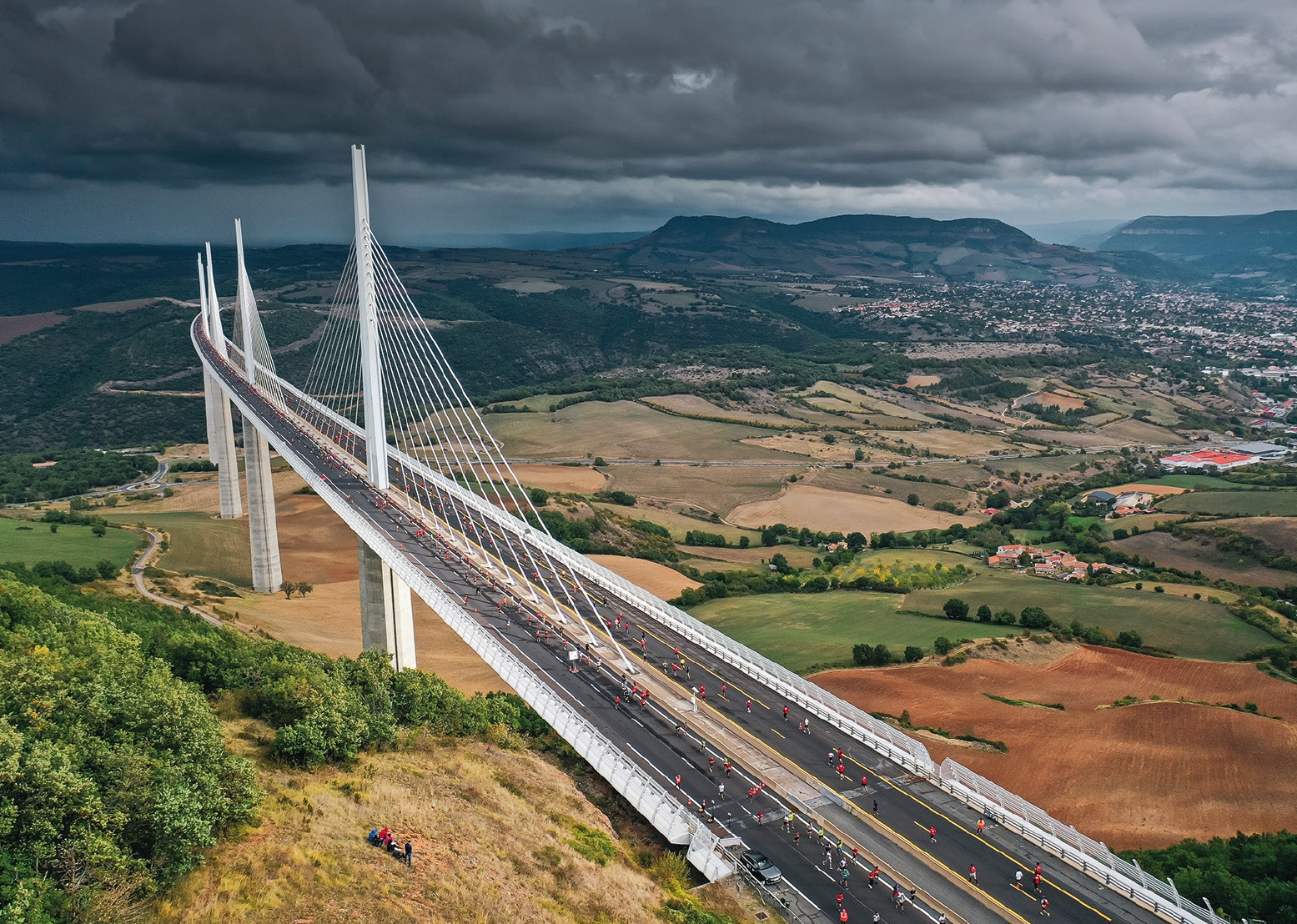 Vue aérienne en plongée du viaduc de Millau. Le ciel est orageux et le pont à haubans blanc se détache nettement sur le paysage.