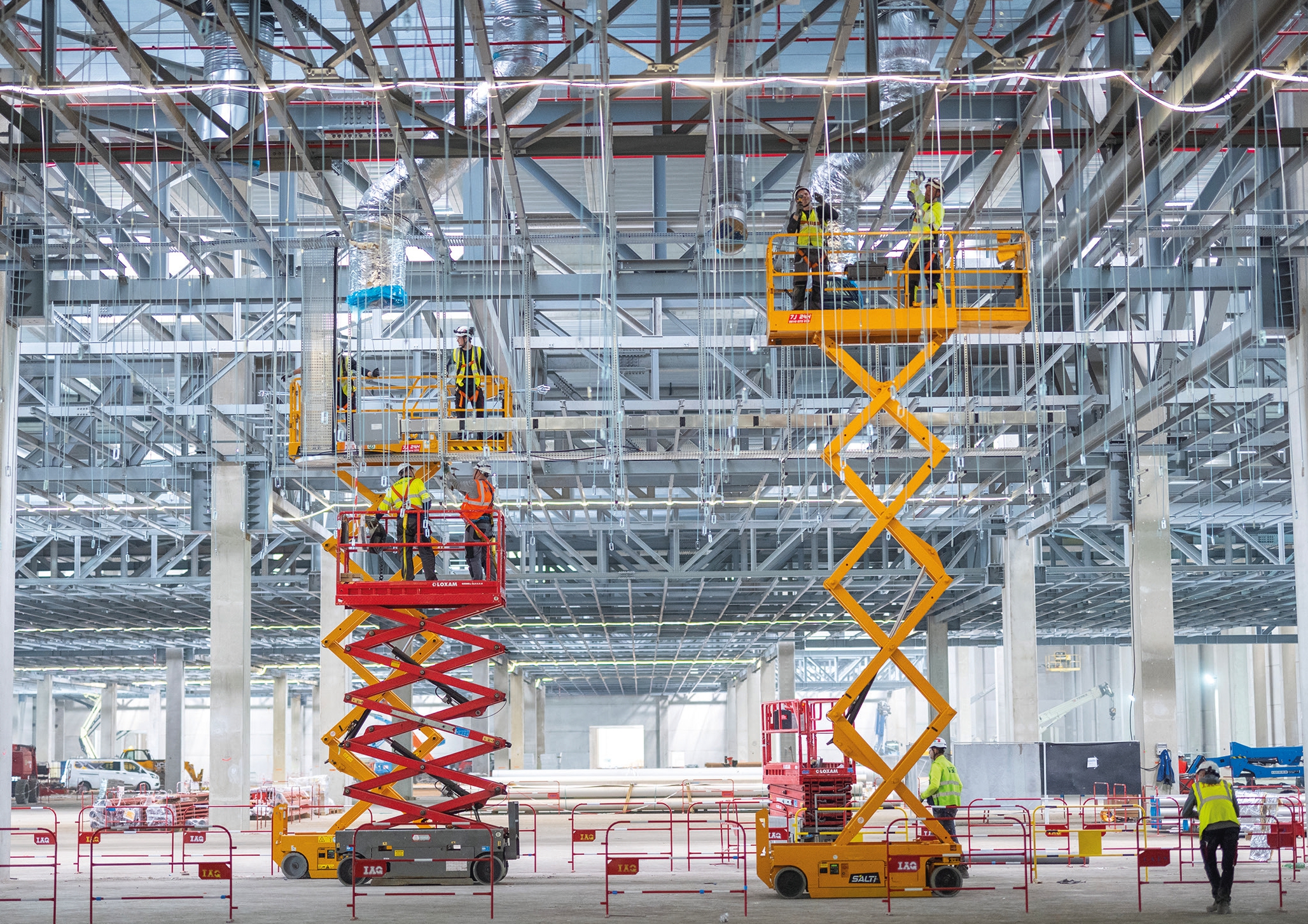 Dans une grande halle en métal très lumineuse, des ouvriers attachent des filins en haut de monte-charges en accordéon jaune et rouge.