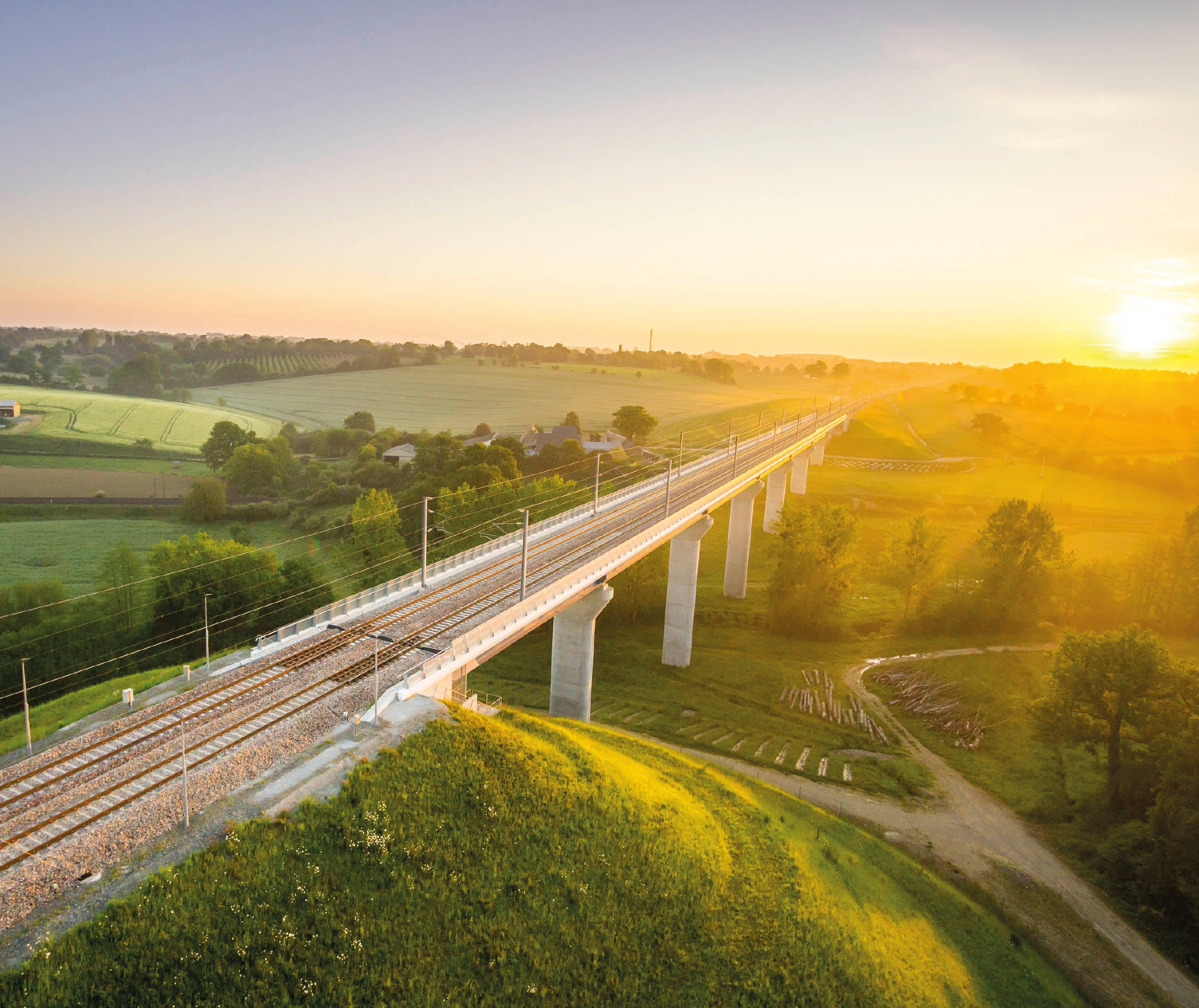 Vue aérienne en plongée d’un pont ferroviaire. Le soleil couchant illumine le paysage.