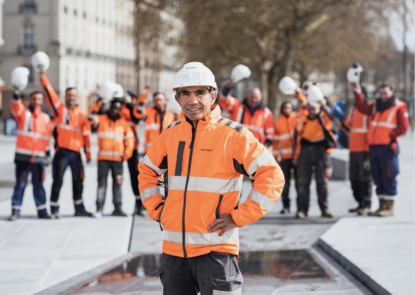 Un ouvrier du B T P avec un casque sourit au premier plan. À l’arrière , on aperçoit d’autres ouvriers qui lèvent leur casque en l’air.